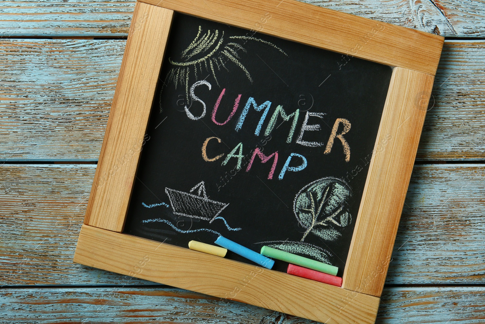 Photo of Blackboard with text SUMMER CAMP, drawings and chalk sticks on wooden background, top view