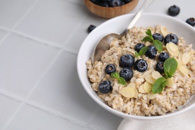 Photo of Tasty oatmeal with blueberries, mint and almond flakes in bowl on white tiled table, closeup. Space for text