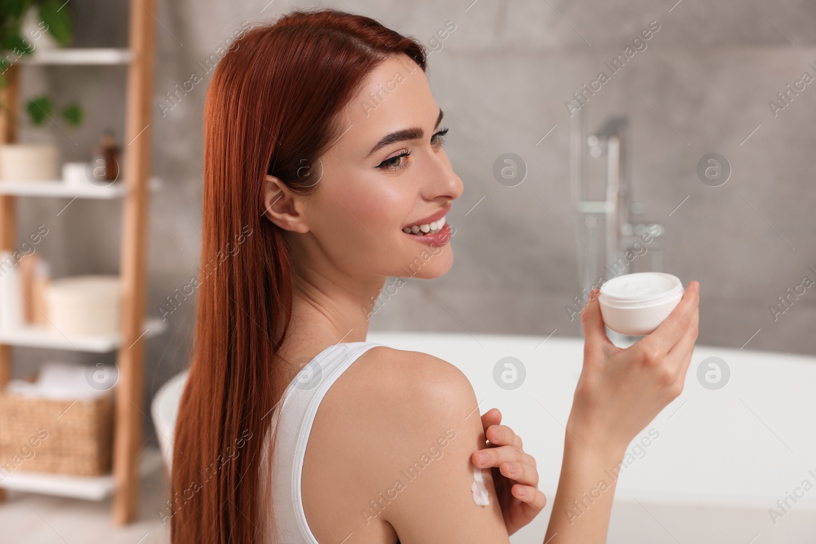 Photo of Beautiful young woman applying body cream onto shoulder in bathroom