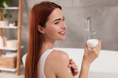 Photo of Beautiful young woman applying body cream onto shoulder in bathroom