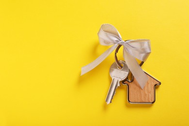 Photo of Key with trinket in shape of house and grey bow on yellow background, top view. Space for text. Housewarming party