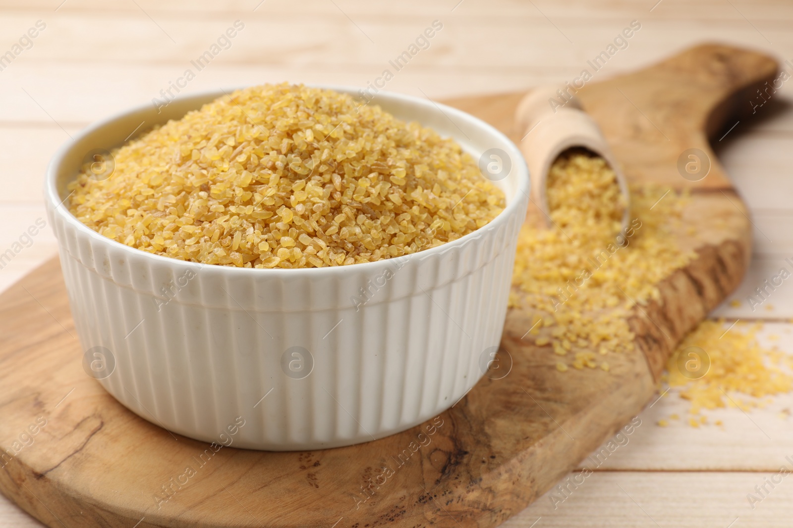 Photo of Bowl and scoop with raw bulgur on light wooden table, closeup