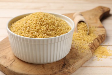 Bowl and scoop with raw bulgur on light wooden table, closeup