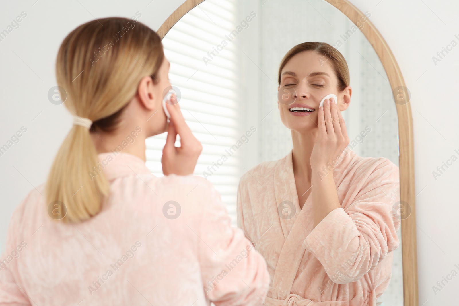 Photo of Beautiful woman removing makeup with cotton pad near mirror indoors