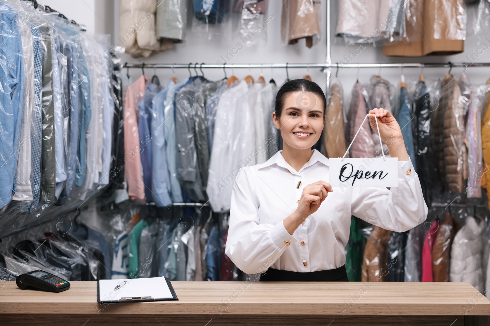 Photo of Dry-cleaning service. Happy worker holding Open sign at counter indoors, space for text