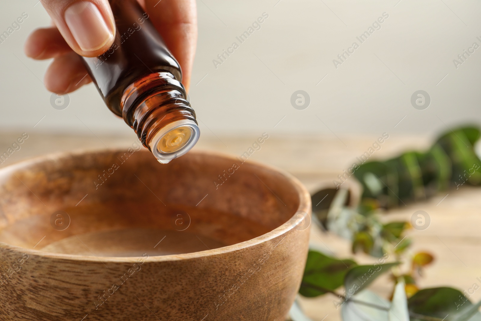 Photo of Woman dripping eucalyptus essential oil from bottle into bowl at wooden table, closeup. Space for text