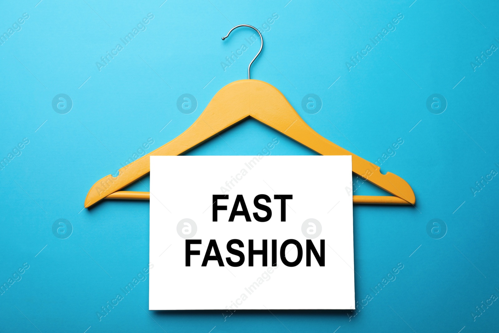 Photo of White card with phrase Fast Fashion and yellow hanger on turquoise background, flat lay