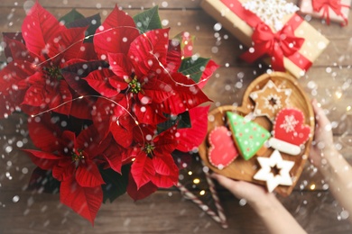 Image of Woman with cookies near traditional Christmas poinsettia flower at wooden table, top view. Snowfall effect