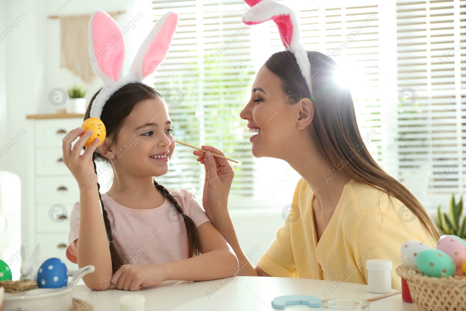 Photo of Happy mother and daughter with bunny ears headbands having fun while painting Easter egg at home