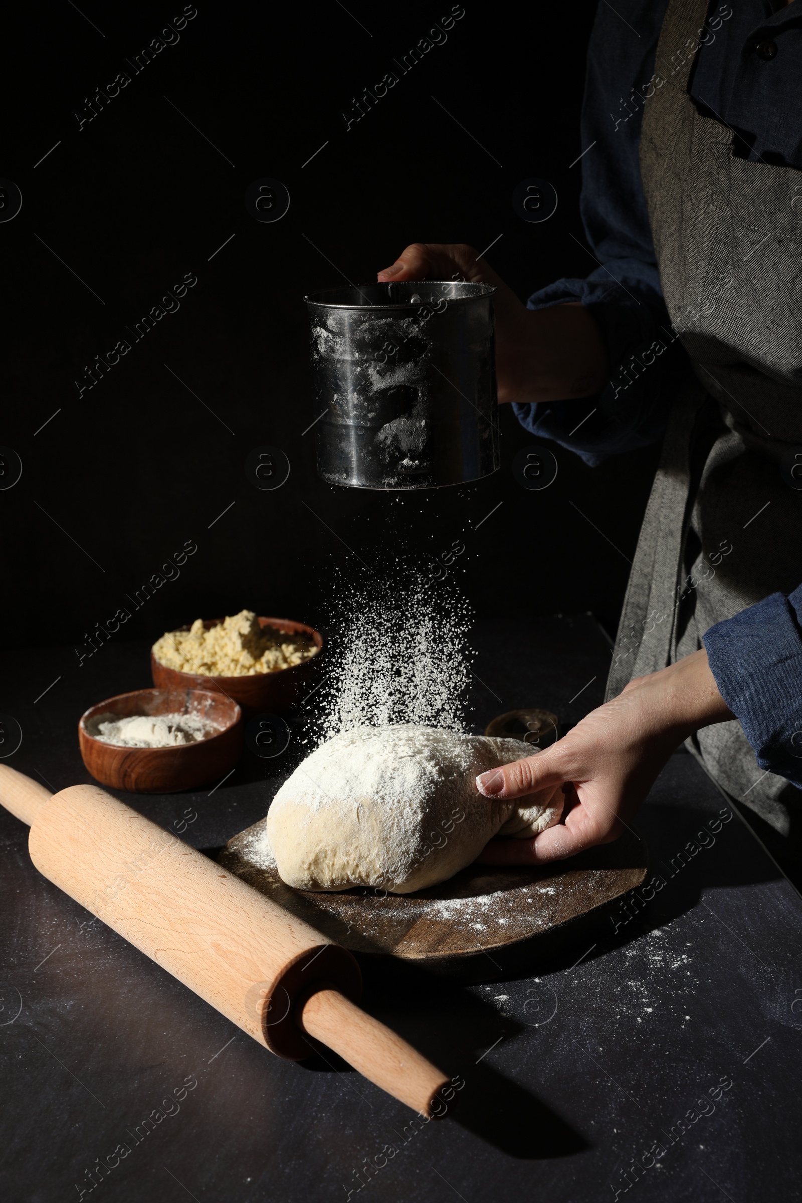 Photo of Woman sprinkling flour over dough at black table, closeup