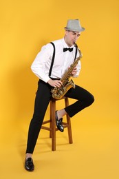 Young man in elegant outfit playing saxophone on yellow background