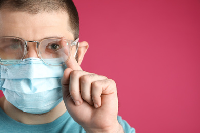 Man wiping foggy glasses caused by wearing disposable mask on pink background, space for text. Protective measure during coronavirus pandemic