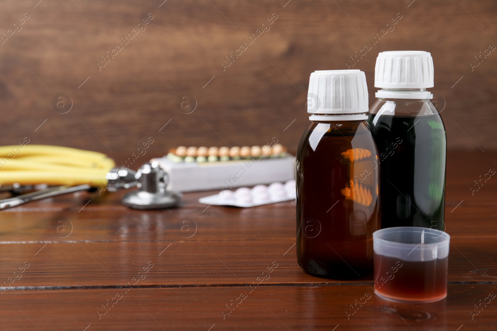 Photo of Bottles of syrup and measuring cup on wooden table, space for text. Cold medicine