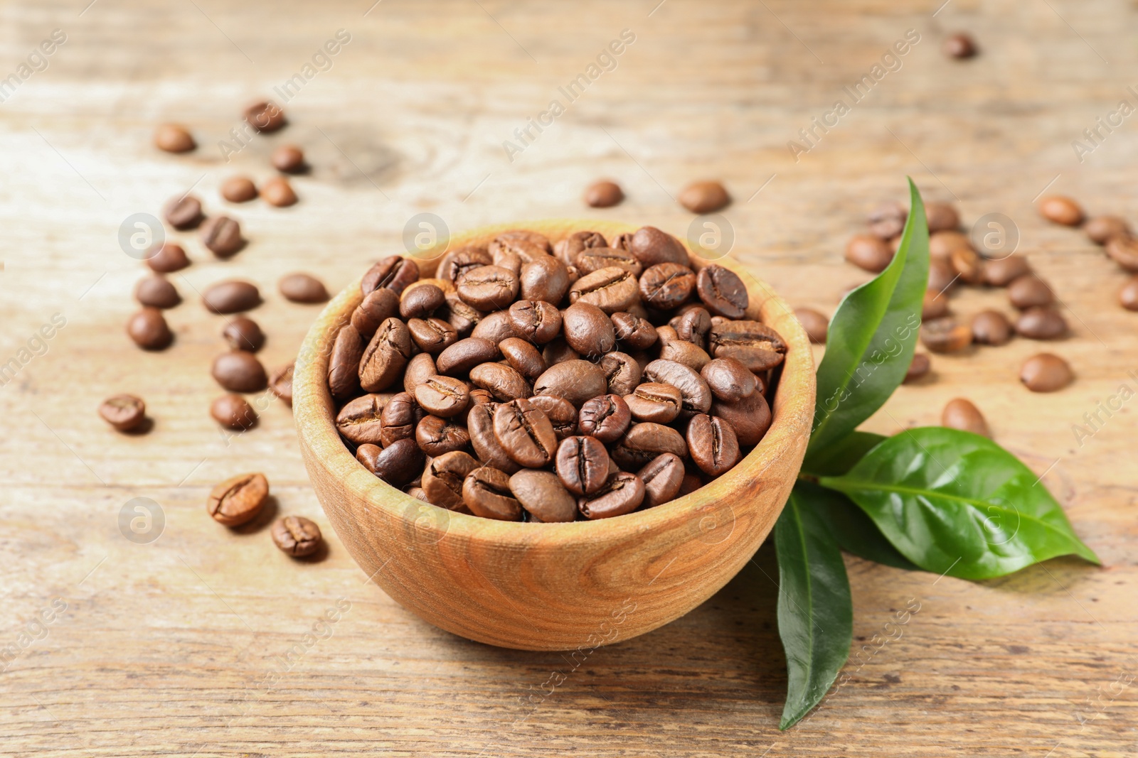 Photo of Bowl of coffee beans and fresh green leaves on wooden table