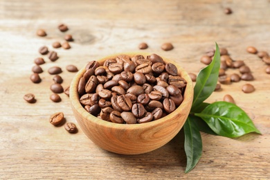 Photo of Bowl of coffee beans and fresh green leaves on wooden table