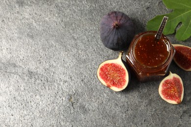 Glass jar with tasty sweet jam, fresh figs and green leaf on grey textured table, flat lay. Space for text