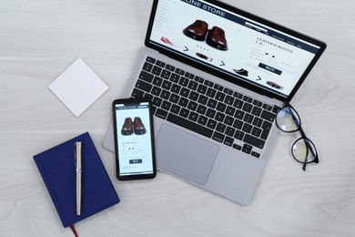 Online store website on laptop screen. Computer, smartphone, stationery and glasses on light grey wooden table, flat lay