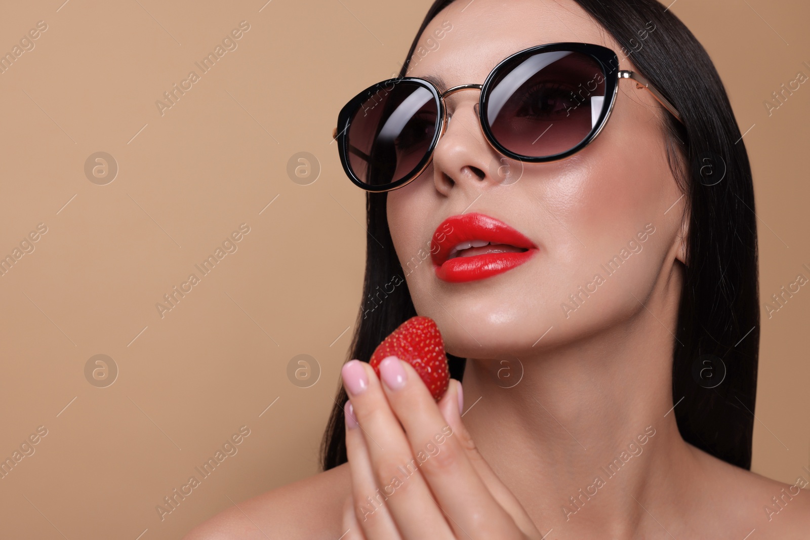 Photo of Attractive woman in fashionable sunglasses holding strawberry against beige background, closeup. Space for text