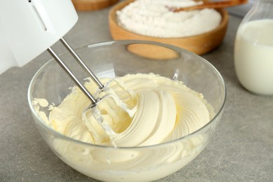 Photo of Whipping white cream in glass bowl with mixer on light grey table, closeup