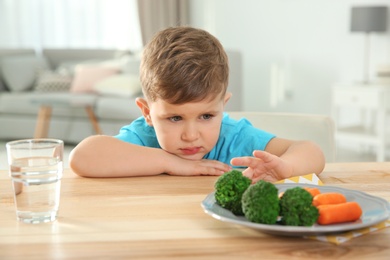 Photo of Unhappy little boy refusing to eat vegetables at table in room