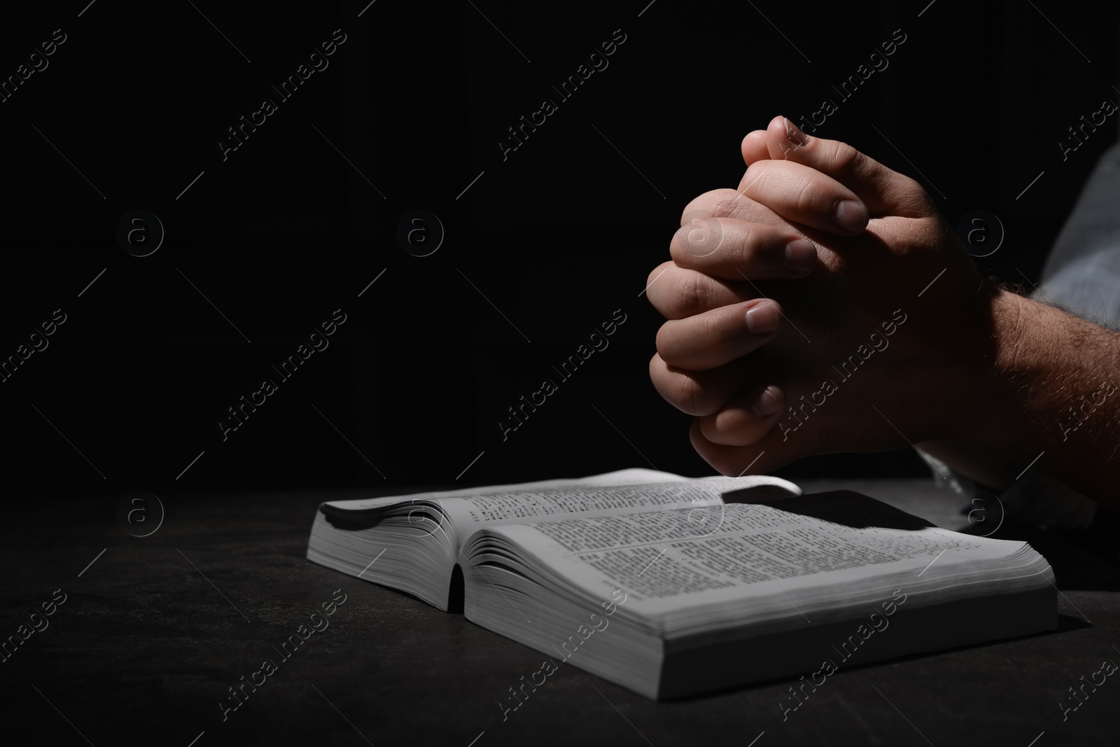 Photo of Religion. Christian man praying over Bible at table against black background, closeup. Space for text