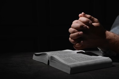 Religion. Christian man praying over Bible at table against black background, closeup. Space for text