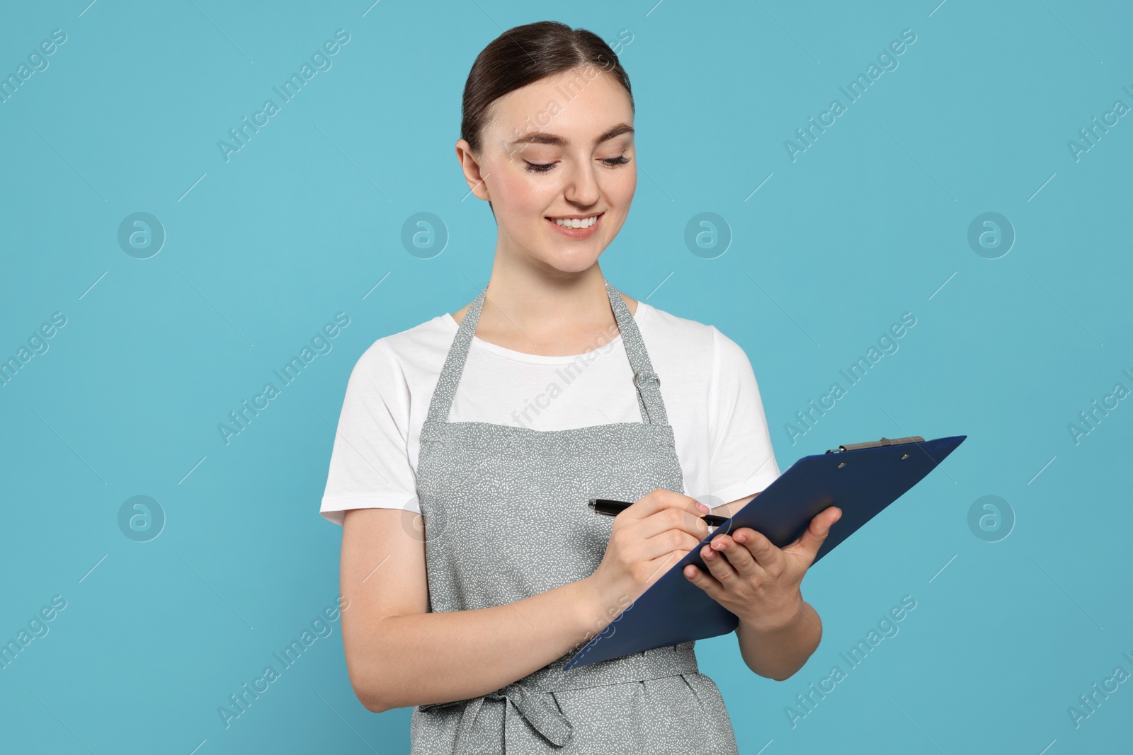 Photo of Beautiful young woman in clean apron with clipboard on light blue background