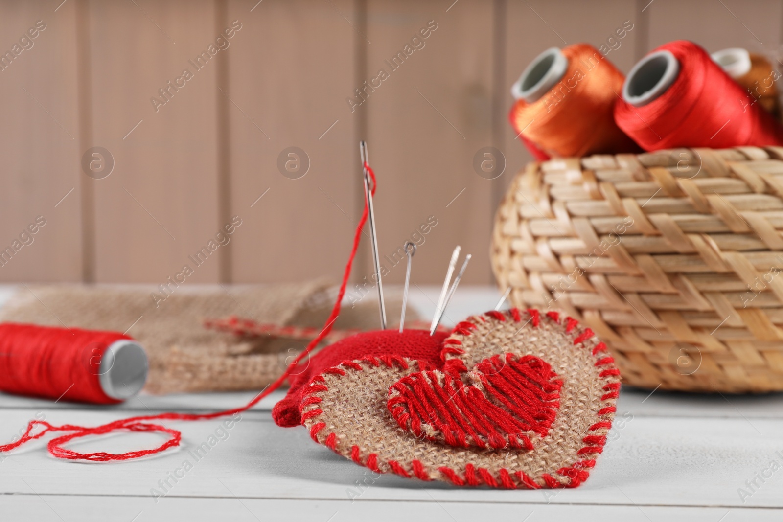 Photo of Heart of burlap fabric with red stitches and spools of threads on white wooden table. Space for text