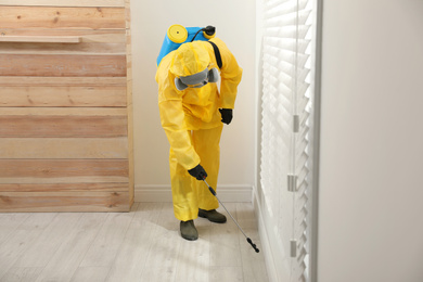 Photo of Pest control worker in protective suit spraying insecticide on floor at home