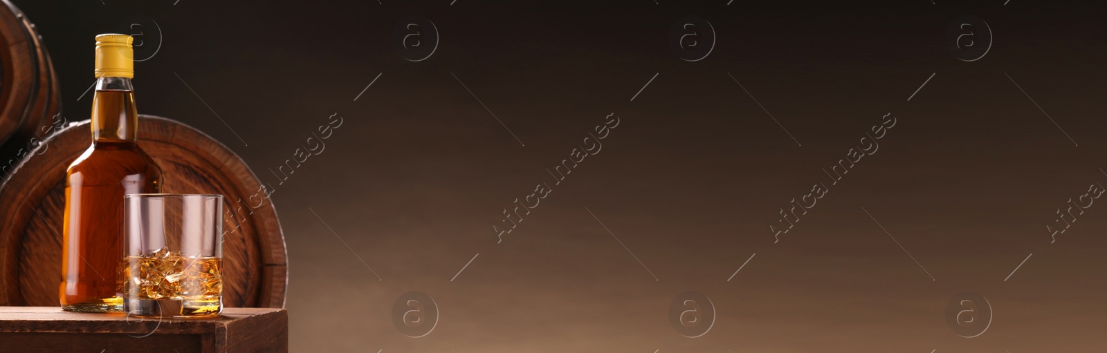 Image of Whiskey with ice cubes in glass and bottle on wooden table near barrels against brown background, space for text. Banner design