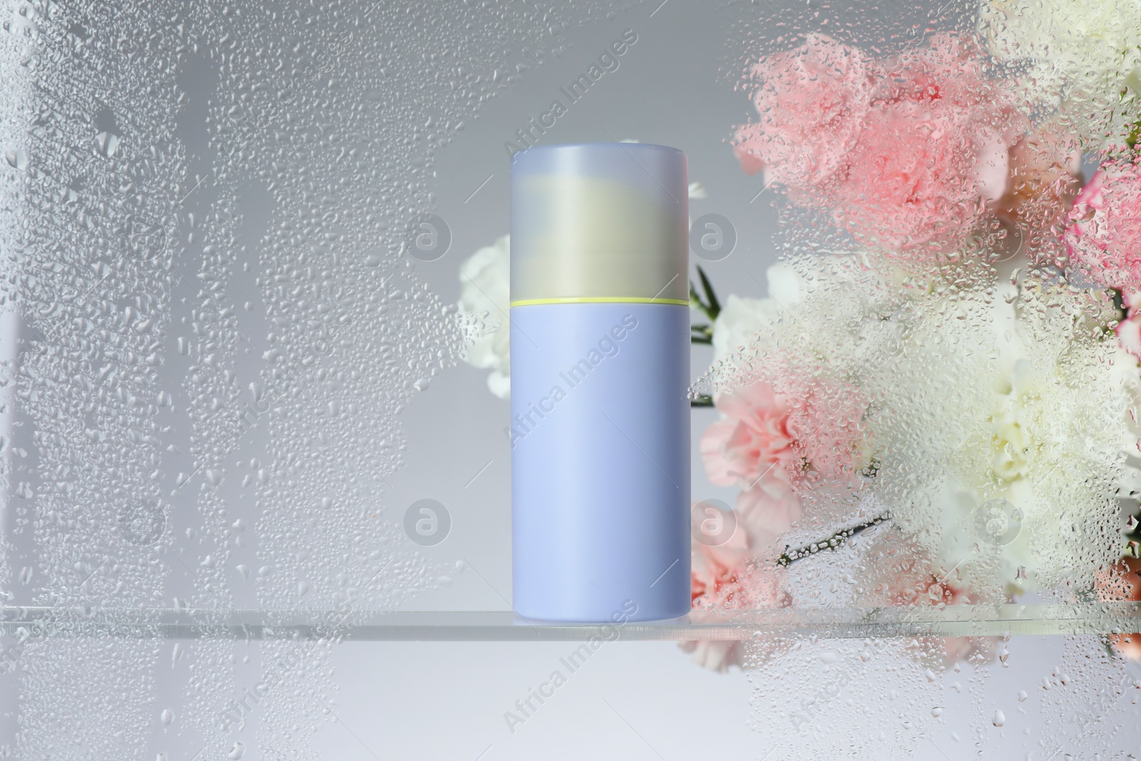 Photo of Bottle with moisturizing cream and beautiful flowers on light background, view through wet glass. Space for text