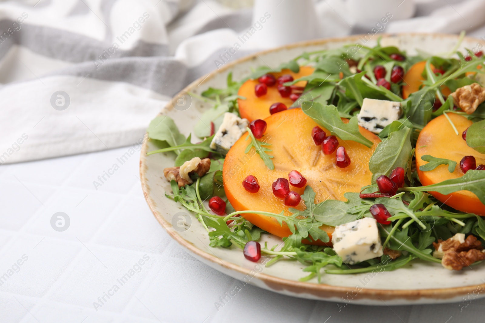 Photo of Tasty salad with persimmon, blue cheese, pomegranate and walnuts served on white tiled table, closeup. Space for text