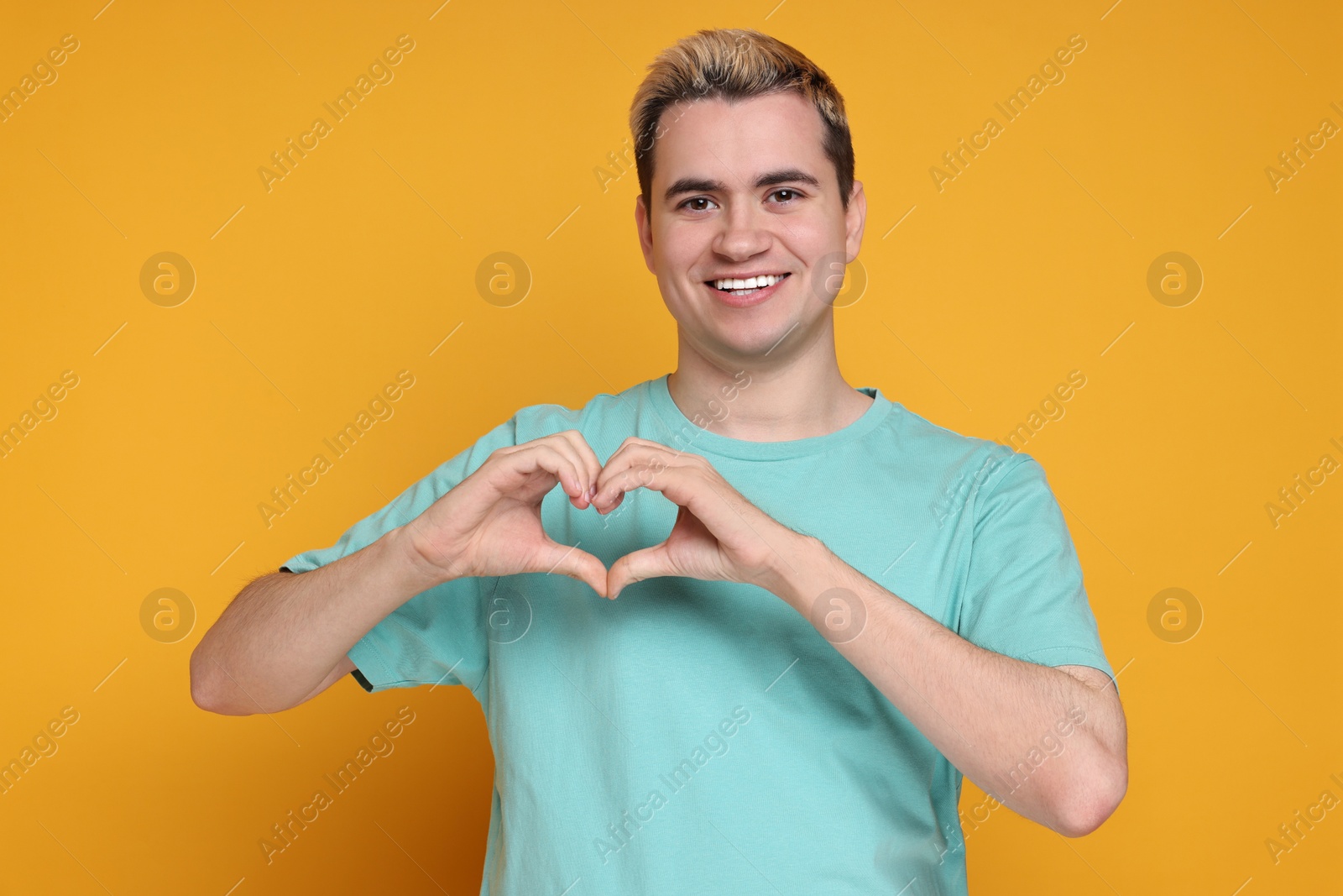 Photo of Young man showing heart gesture with hands on orange background