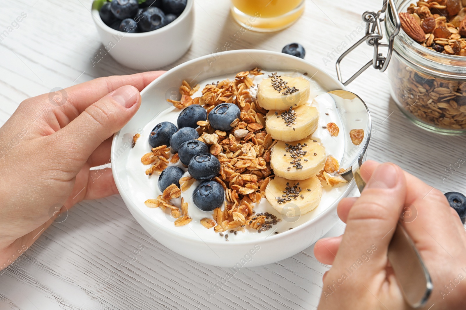 Photo of Woman eating tasty yogurt with oatmeal, banana and blueberries at white wooden table, closeup