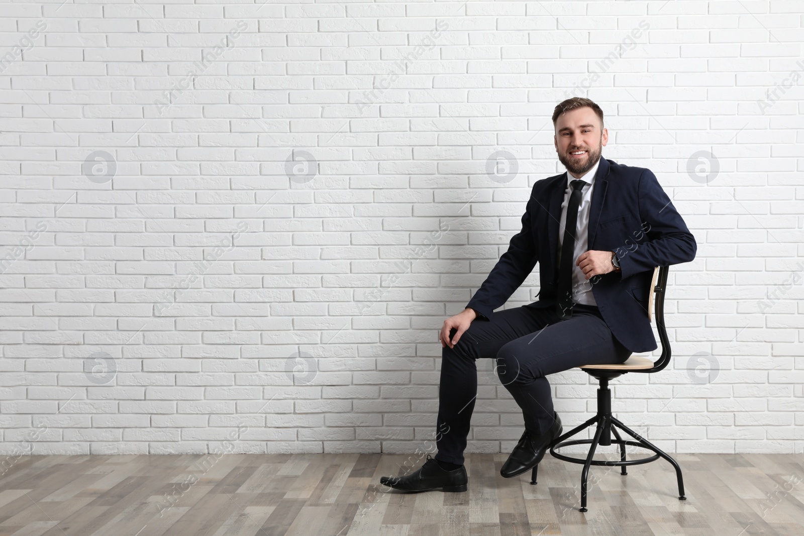 Photo of Young man sitting in office chair near white brick wall indoors, space for text