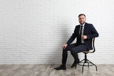 Young man sitting in office chair near white brick wall indoors, space for text