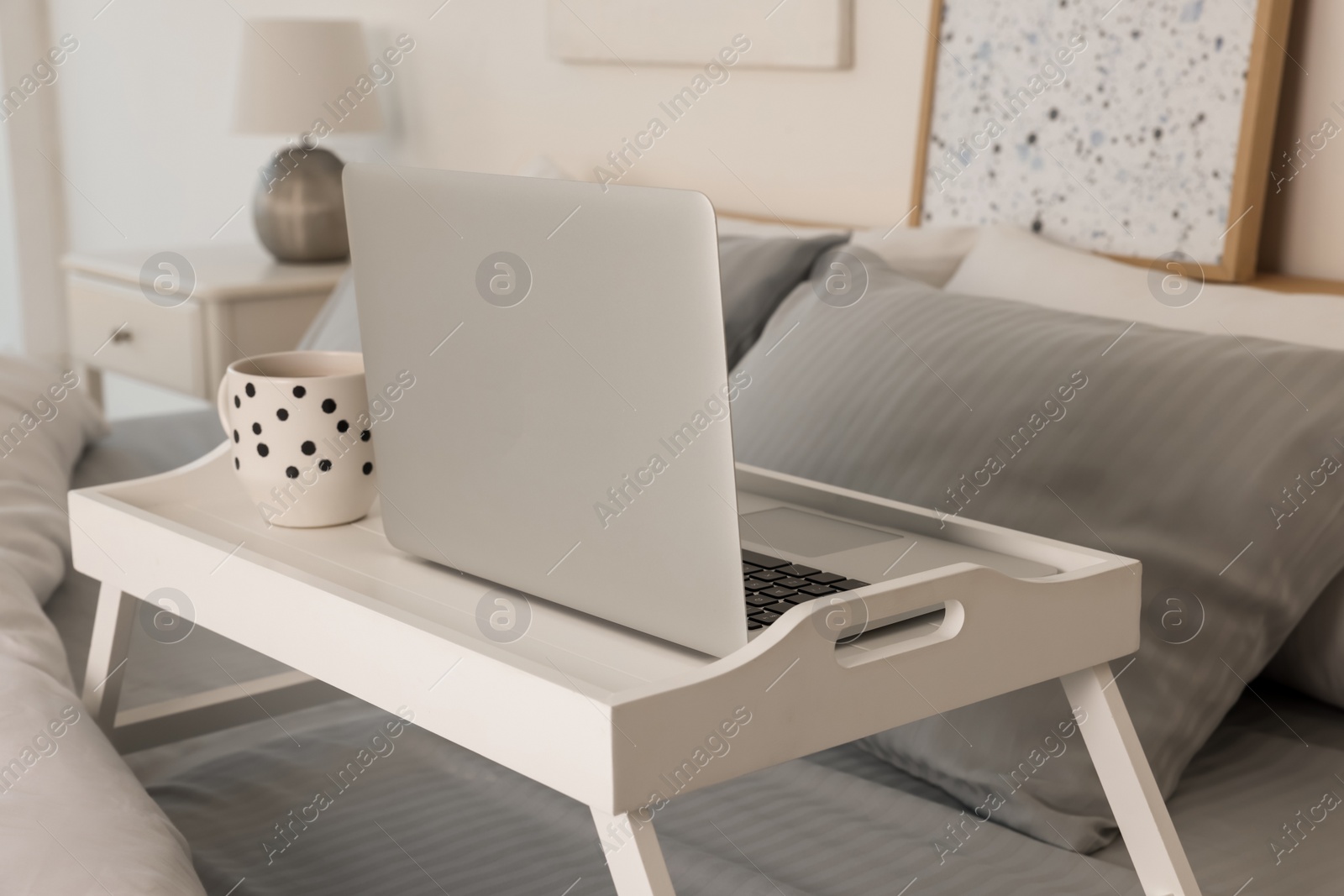 Photo of White tray table with laptop and cup of drink on bed indoors