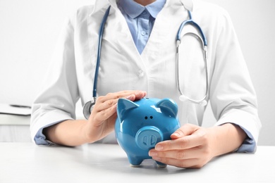 Doctor with piggybank at white table indoors, closeup. Medical insurance concept