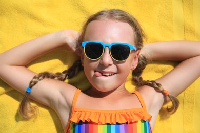 Photo of Happy little girl in sunglasses lying on beach towel outdoors, top view