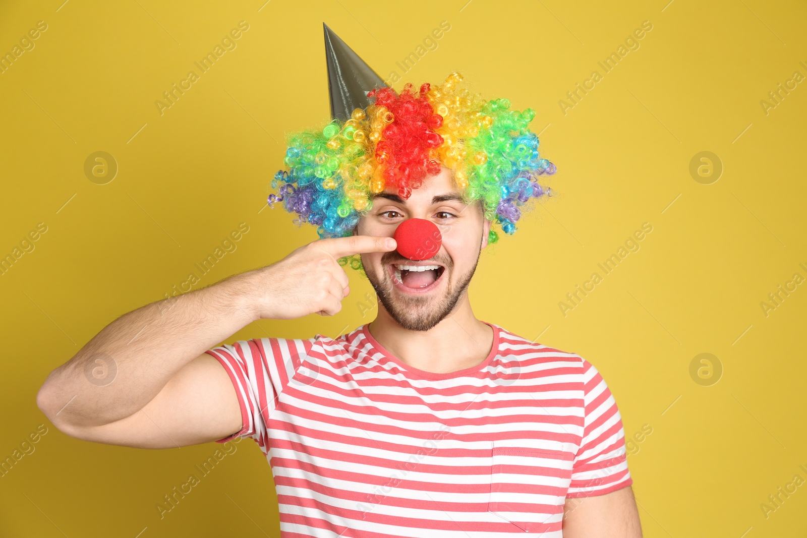 Photo of Emotional young man with party hat and clown wig on yellow background. April fool's day