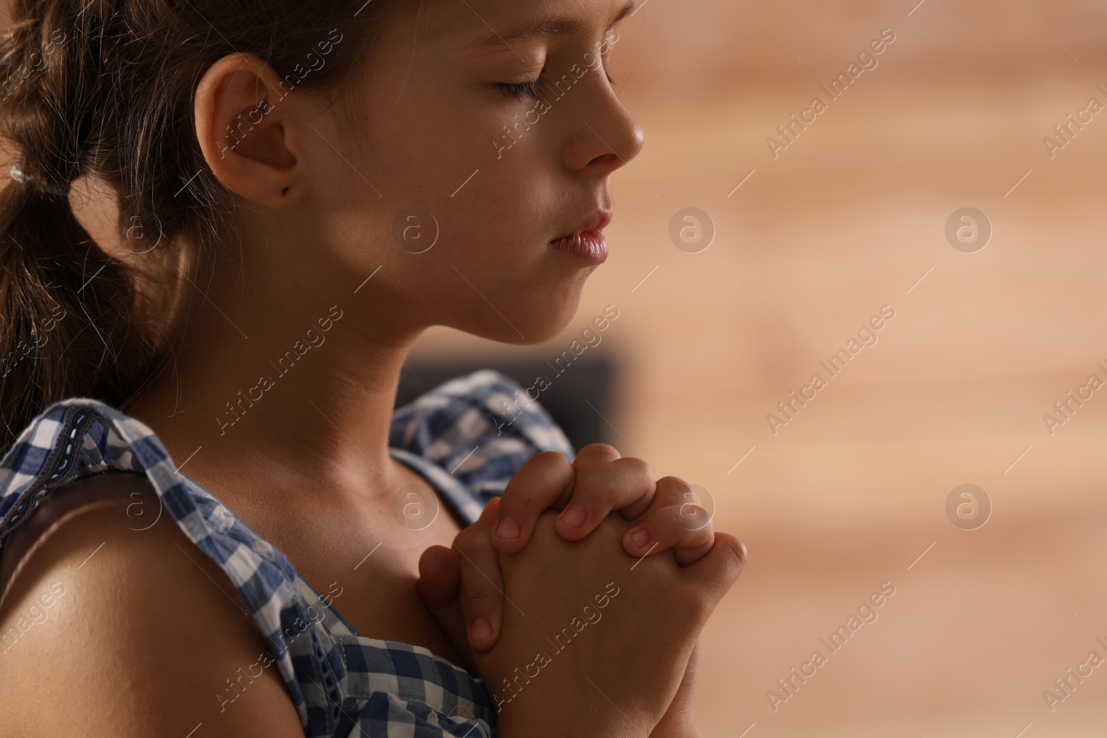 Photo of Cute little girl with hands clasped together praying on blurred background, closeup. Space for text