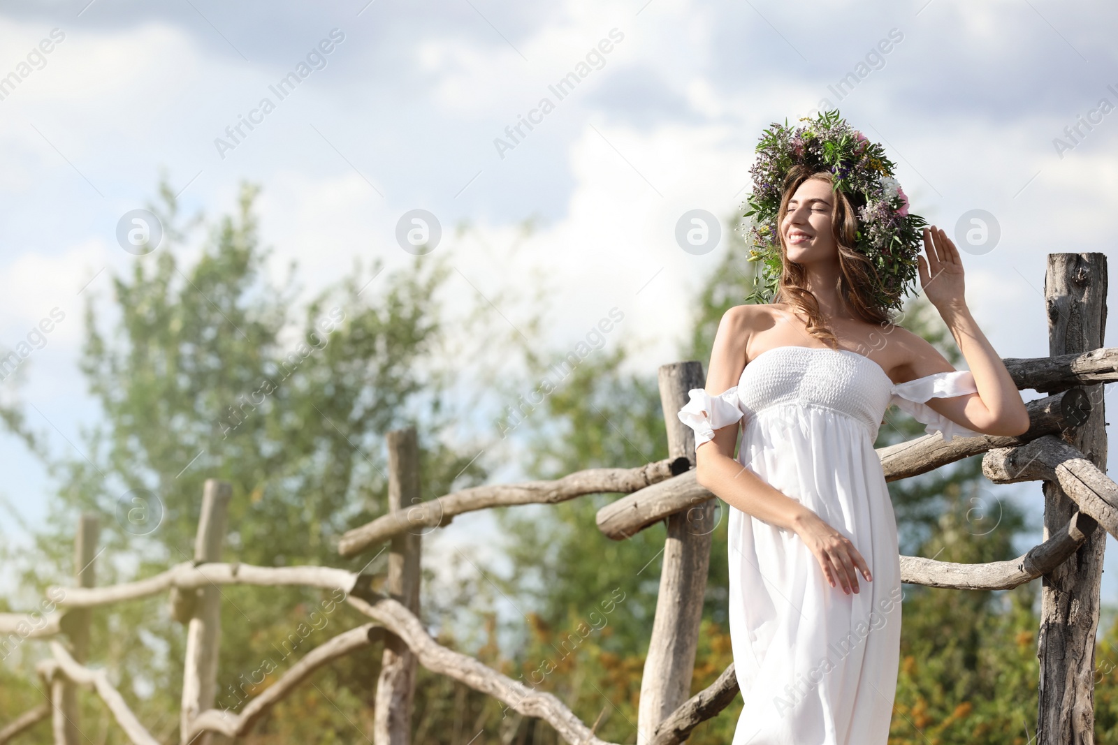 Photo of Young woman wearing wreath made of beautiful flowers near wooden fence on sunny day