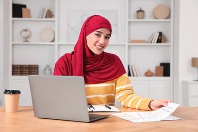 Muslim woman writing notes near laptop at wooden table at home