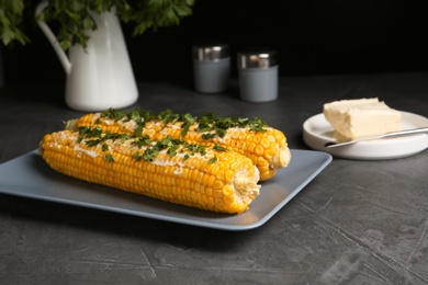 Delicious boiled corn cobs with butter and parsley on gray table against black background