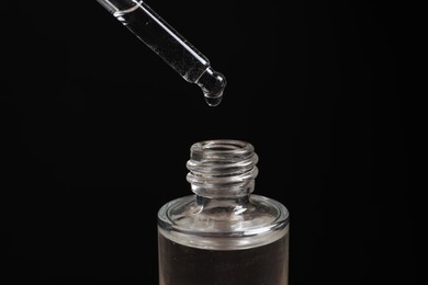 Photo of Dripping cosmetic serum from pipette into bottle on black background, closeup