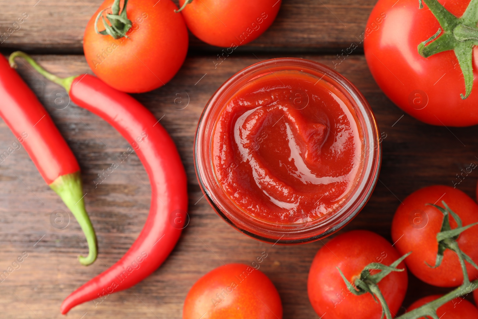 Photo of Jar of tasty ketchup, chili peppers and tomatoes on wooden table, flat lay