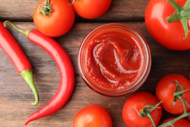 Jar of tasty ketchup, chili peppers and tomatoes on wooden table, flat lay