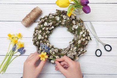 Woman decorating willow wreath with daffodil and hyacinth flowers at white wooden table, top view