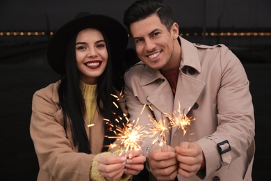 Photo of Couple in warm clothes holding burning sparklers near building