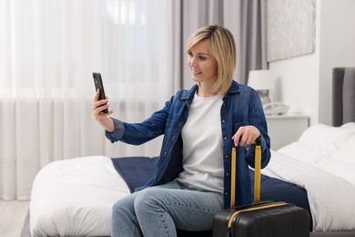 Photo of Smiling guest with suitcase taking selfie on bed in stylish hotel room
